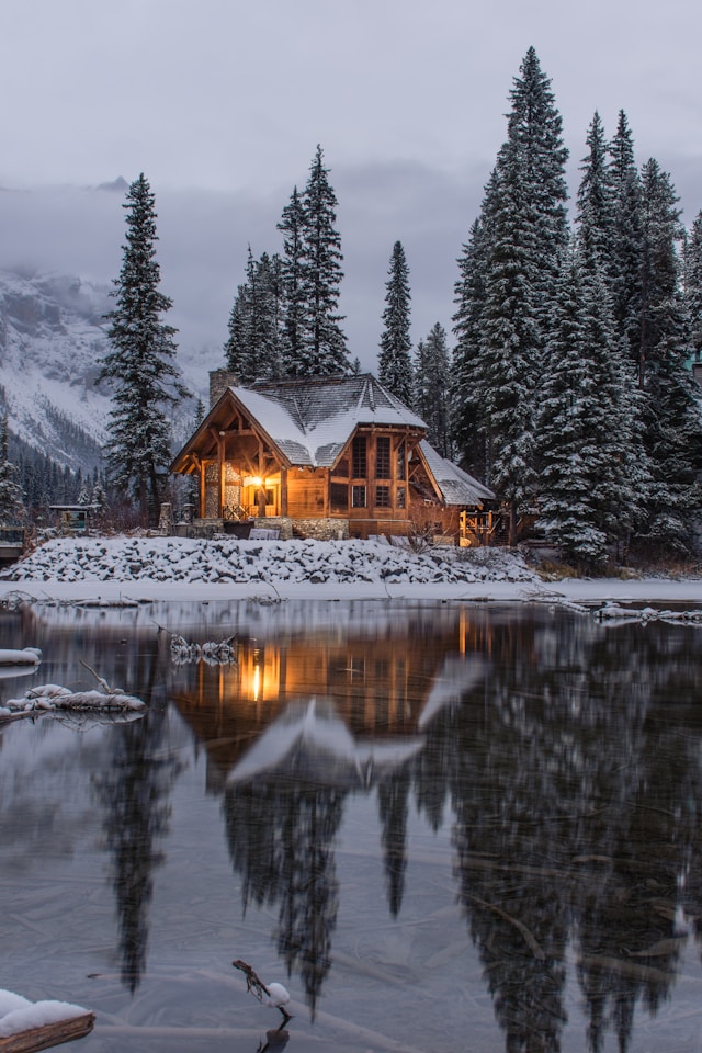 House made of wood on a snow clad in front of a frozen lake surrounded by fern trees in front of mountains