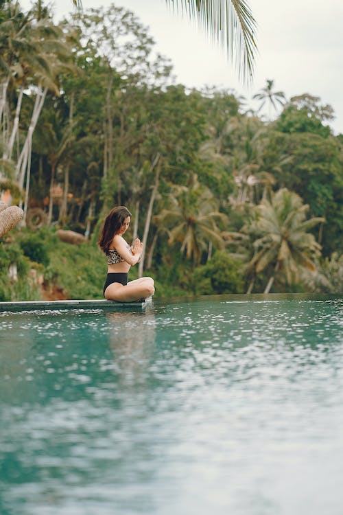 Free Side view full length of slim unrecognizable brunette in swimwear sitting in lotus pose and meditating while spending time near peaceful pond in tropical country Stock Photo