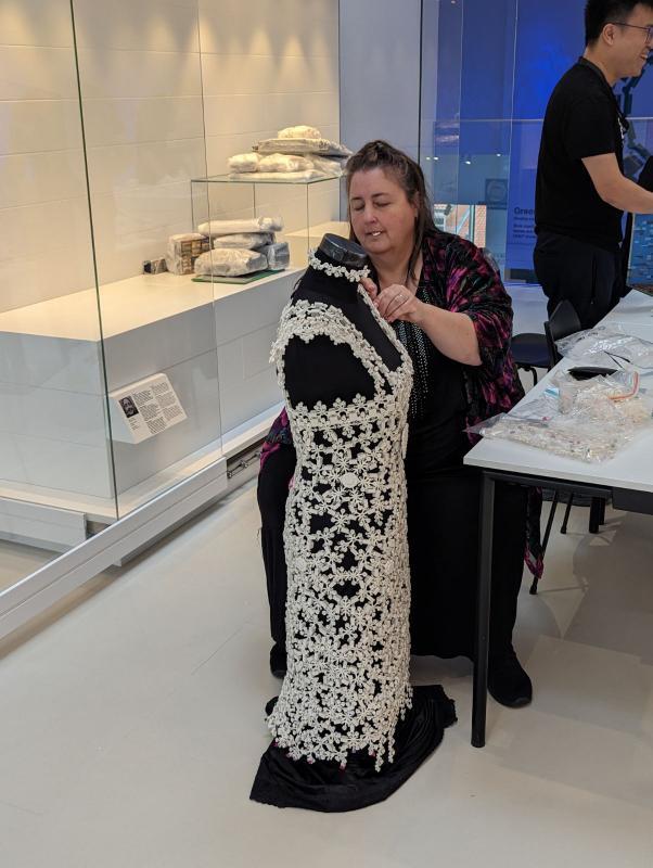 A photo of Veronica Young in the LEGO Masterpiece Gallery, sitting while assembling part of her LEGO wedding dress, which is on a black mannequin
