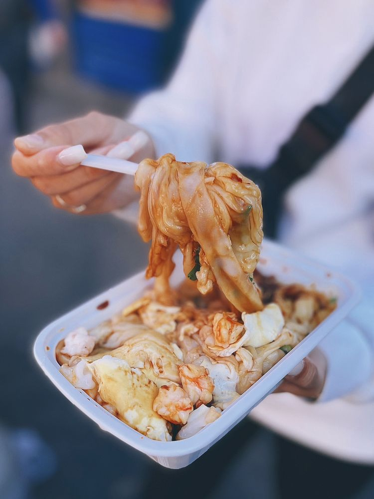 Photo from Cheong Fun Cart's Yelp page. A nicely manicured hand holds a fork. On the end of the fork is a thick twirl of cheong fun noodles. The other hand holds the rest of the takeout container holding more noodles.