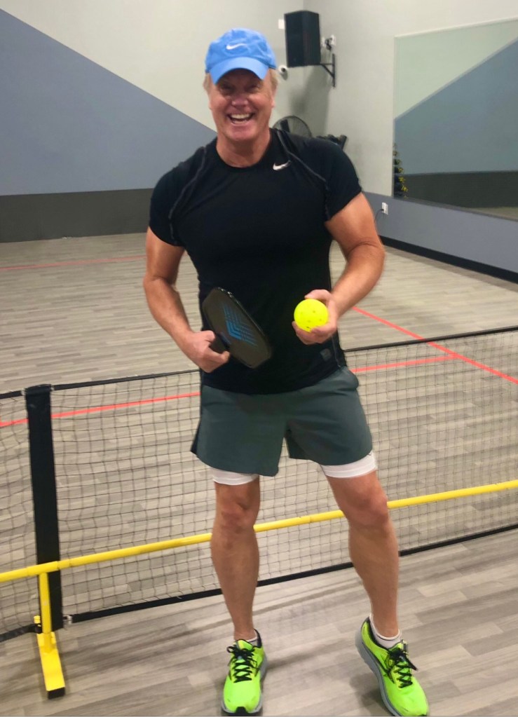 Rick Domeier smiles during a game of tennis, a racket and ball in each hand.
