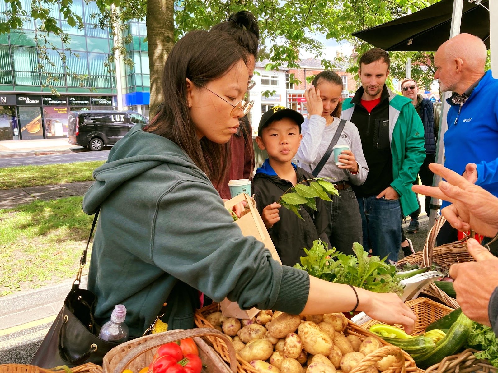 A woman selects vegetables from a basket on the market stall, while other shoppers in the queue look on. 