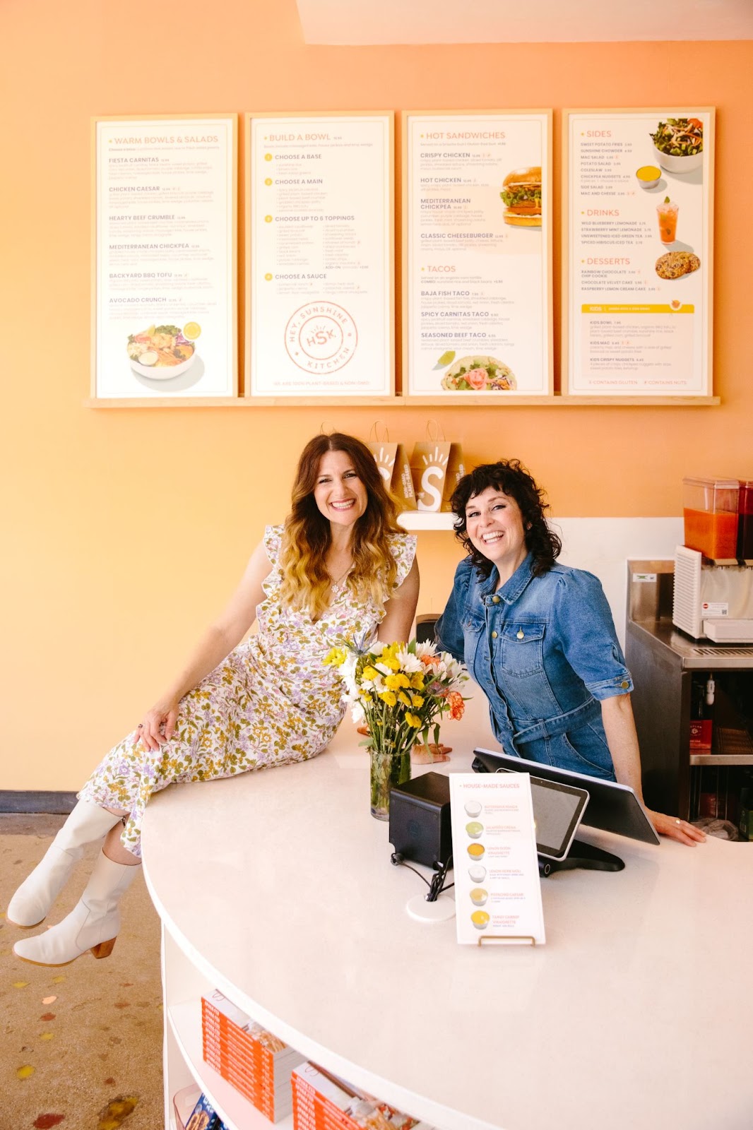 Hey Sunshine Kitchen employees smile at the checkout counter, large menus are hanging on an orange wall