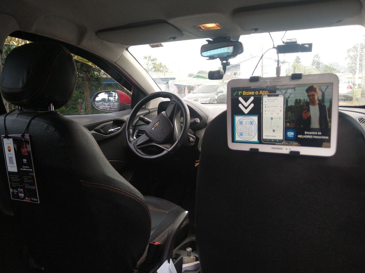 A imagem mostra o interior de um carro, provavelmente um serviço de transporte por aplicativo, visto pelo ângulo do banco de trás. O painel e o volante estão à frente, e há um tablet montado no encosto do banco do passageiro da frente, mostrando uma propaganda com um QR code e a instrução "Baixe o App", além de um homem sorridente usando óculos de sol. Há também um identificador de motorista pendurado no encosto do banco, mas os detalhes específicos não são visíveis. Através da janela lateral, pode-se ver o que parece ser um estacionamento ou uma rua com árvores e parte de outro veículo.





