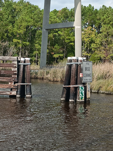 In the background there is a high-rise bridge and the dolphins (fenders) that protect the bridge.  In the foreground, pilings rise out of the water and there are numbers indicating how many feet of clearance are available for boats going under the bridge.  The numbers indicate there is only 64.5 feet of clearance.