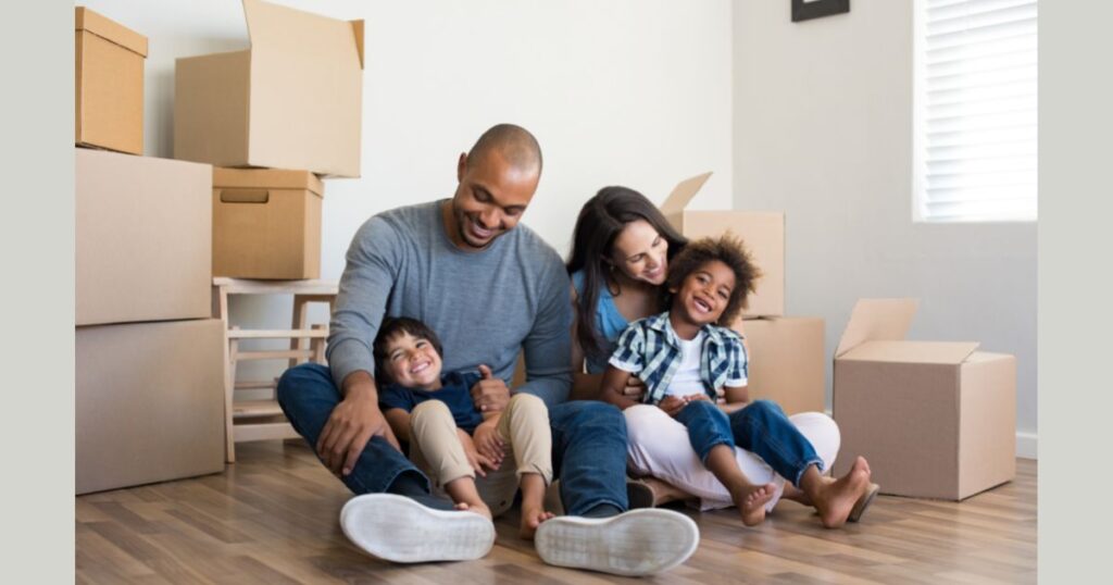A mother and father sit on the floor with their children in their laps with moving boxes in the background.