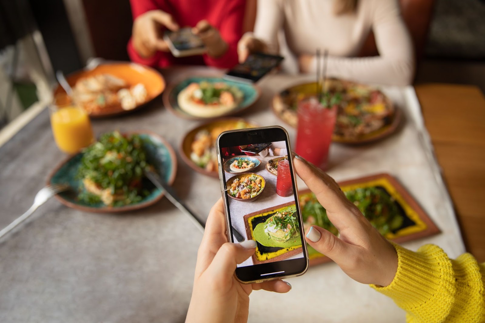 Mãos de uma mulher tirando foto de pratos de comida em uma mesa de restaurante