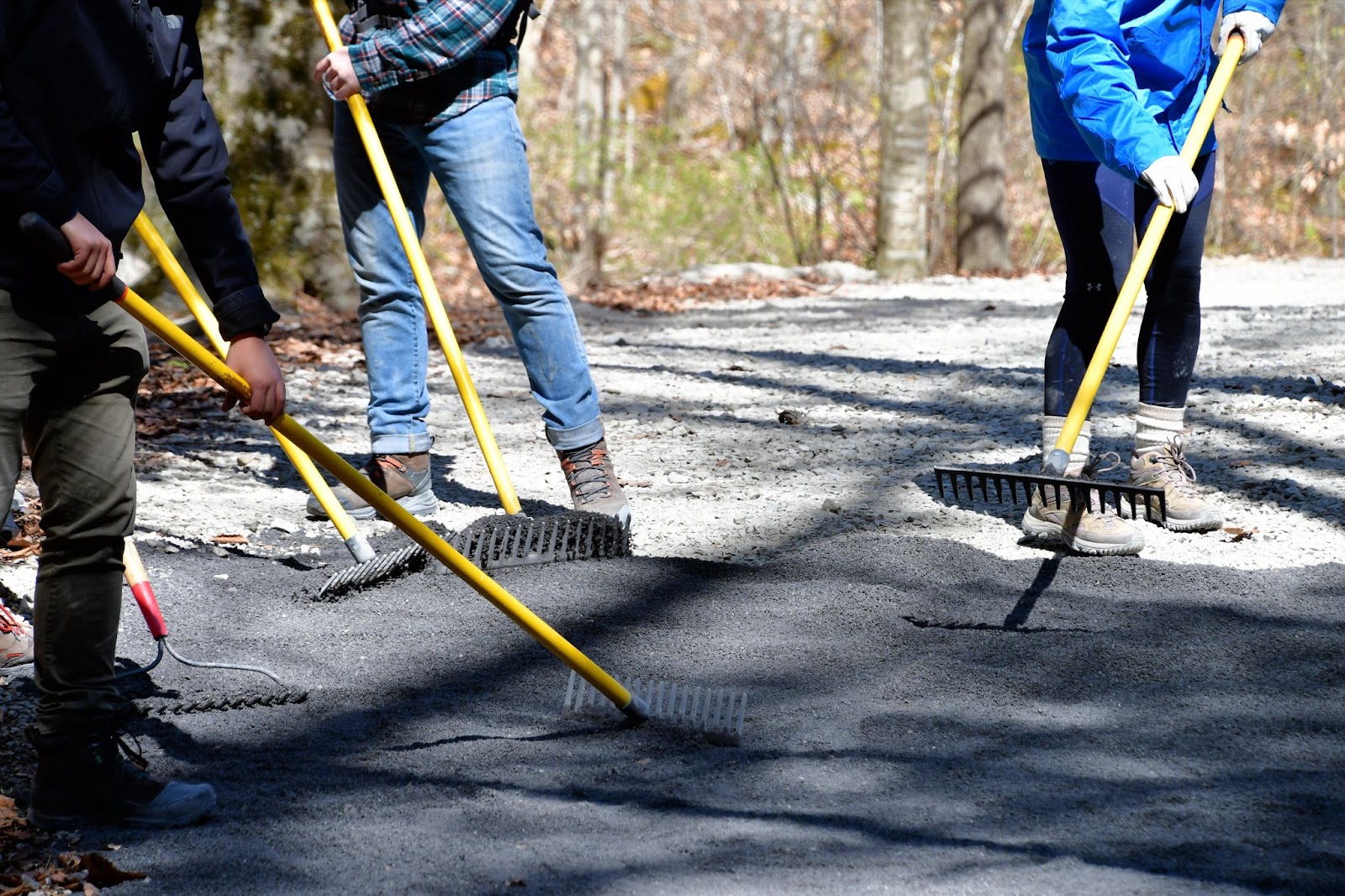 Three workers using rakes, shovels, and other hand tools to spread gravel on outdoor asphalt pathways.