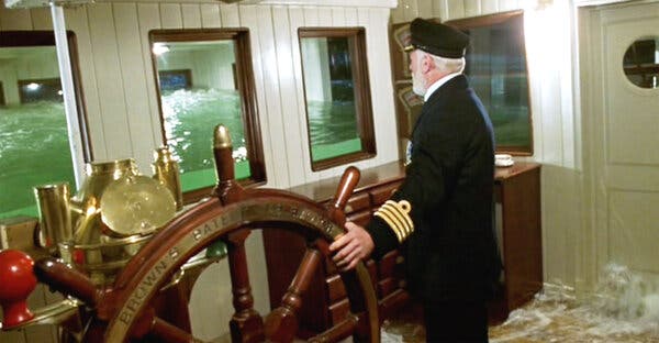 A man in a captain’s uniform stands at the wheel of a ship, looking through a window as the room in front fills with water.