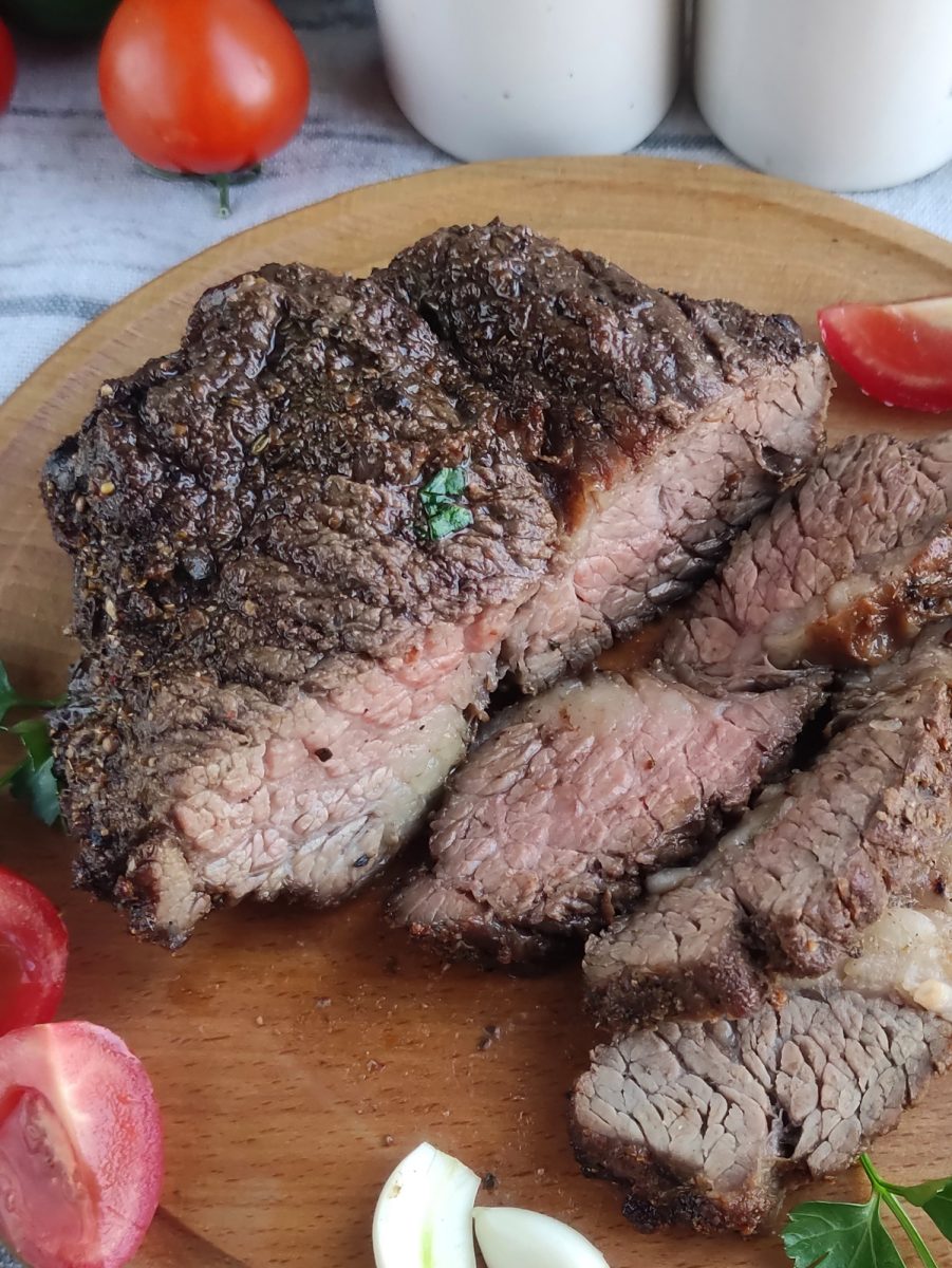 Air fryer ribeye steak on a wooden cutting board with tomatoes and herbs.