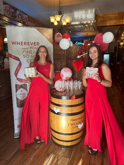 Two women in red dresses standing next to a barrel of wine glasses

Description automatically generated