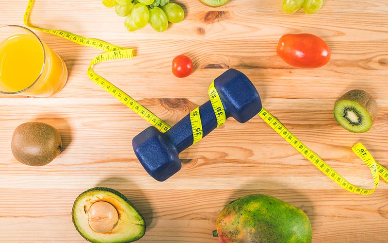 Yellow measuring tape wrapped around a blue dumbbell next to avocados and other fresh fruit