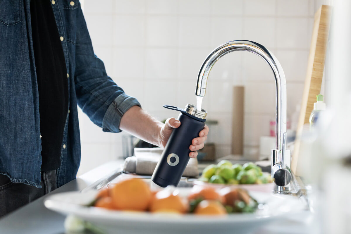 person filling up water bottle at kitchen faucet with bowl of fruit in foreground