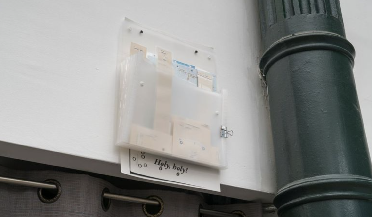 Image: Installation view of Untitled and unlisted work by olivier at Roots & Culture Contemporary Art Center. An open transparent folder with a variety of notes and a paper behind it reading "Holy, holy!" with various o's on it hung on the ceiling next to a pipe and above a curtain. Photography by Robert Chase Heishman.