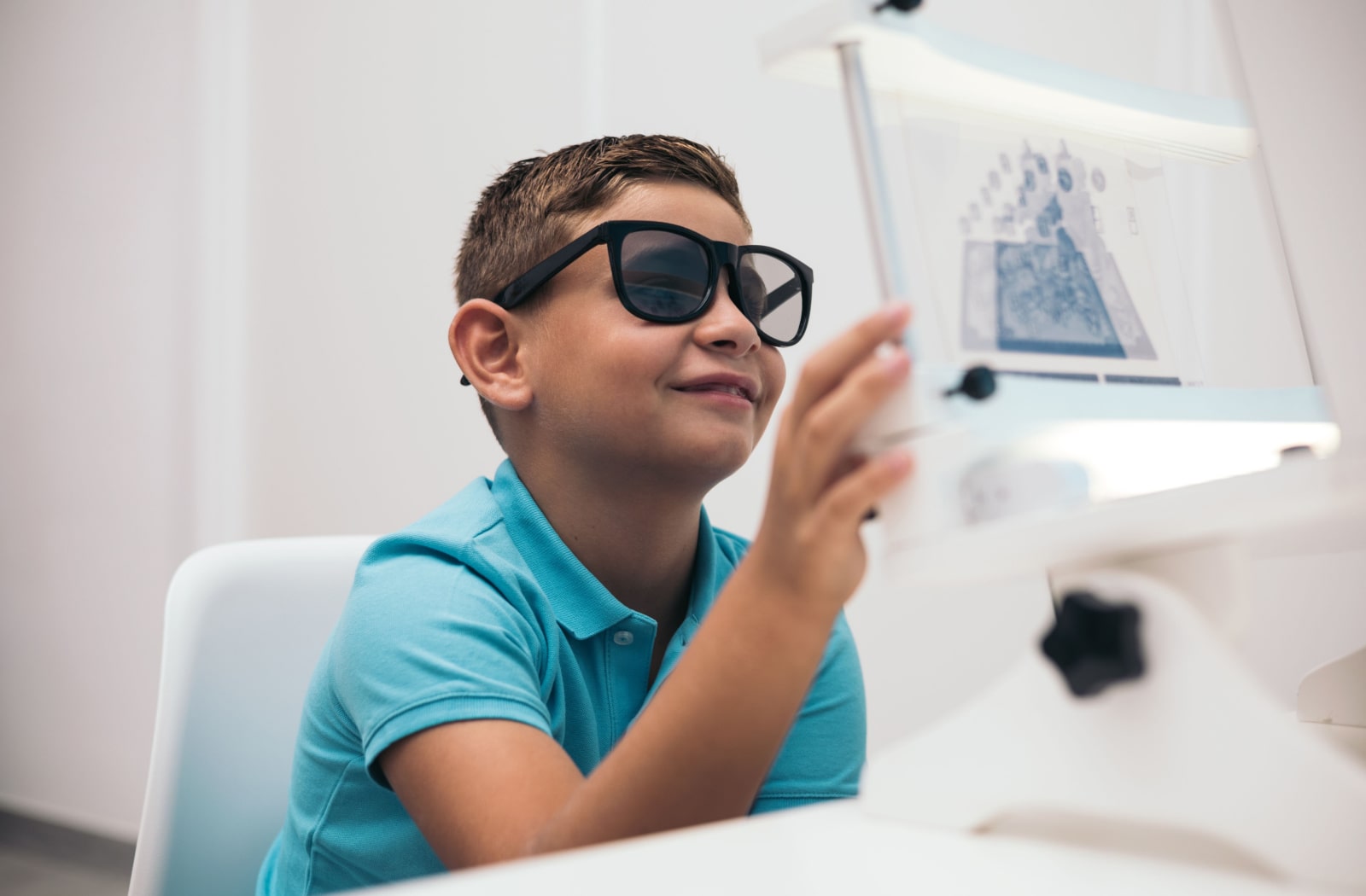 A child is undergoing a convergence/divergence test using vectograms. The image shows the child wearing polarized glasses and looking at a series of images on a board. The vectograms appear to have overlapping shapes in different colours and sizes, designed to test the child's ability to perceive depth and focus on objects at different distances.