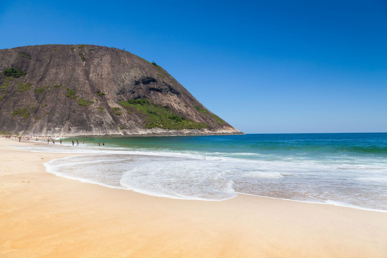 Praia com mar azul sem ondas chegando a areia dourada. Grande rocha ao lado esquerdo da foto.