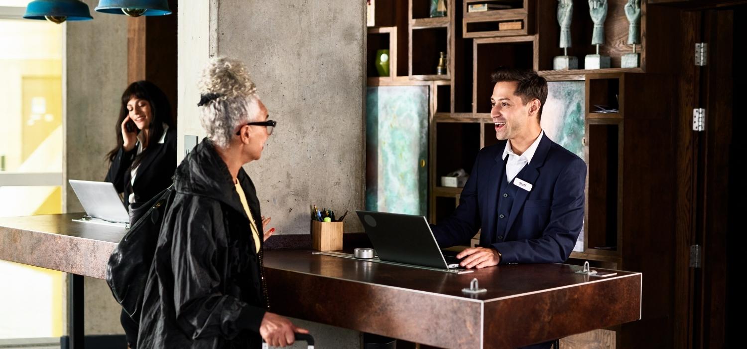 A receptionist greeted the hotel guest with a smile wearing a blue suit and working on his laptop. 