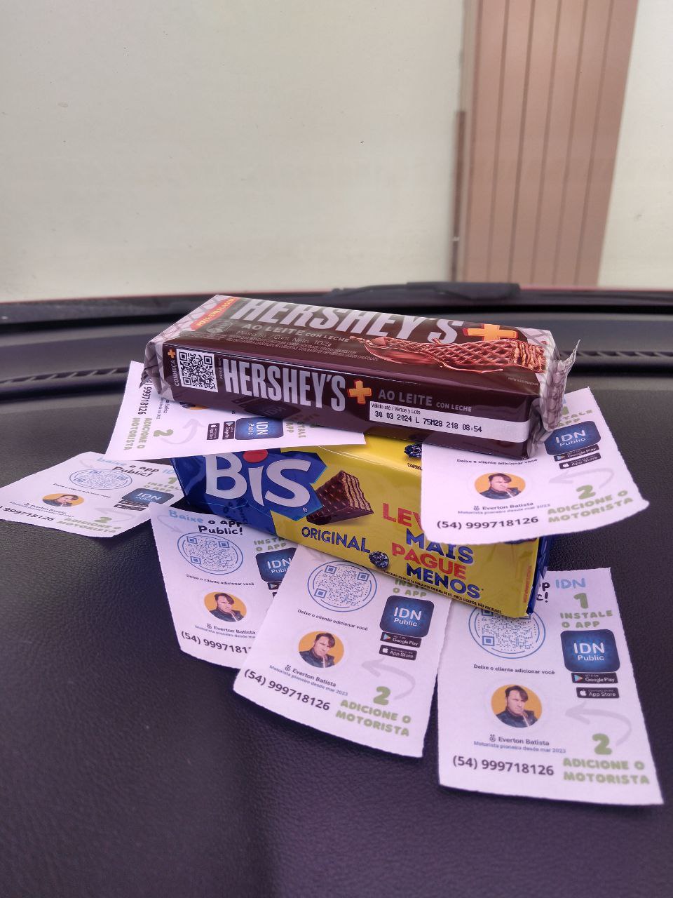 A imagem exibe um painel de carro sobre o qual estão dispostos alguns itens. Há uma barra de chocolate Hershey's sabor ao leite e uma caixa de chocolates Bis. Além dos chocolates, há diversos cartões com QR Codes e informações de contato de motoristas de um serviço de transporte privado, indicando uma promoção ou meio de adicionar motoristas ao aplicativo de serviço. Os cartões apresentam fotos dos motoristas, seus nomes e números de telefone, junto com a logo de um aplicativo chamado "IDN". A fotografia tem um fundo desfocado, onde se pode perceber uma parede e uma porta ao fundo, indicando que o carro está estacionado próximo a um edifício.





