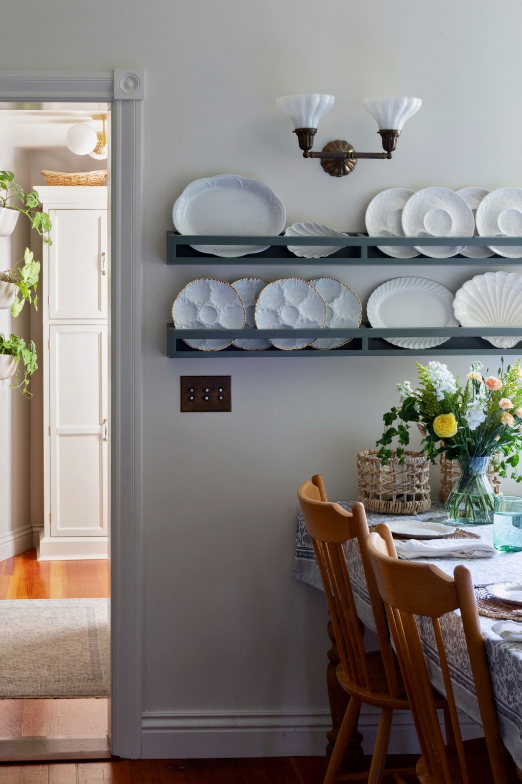 Wooden chairs surround a rectangular table covered with a floral tablecloth, showcasing white ceramic plates on open shelves in Kim Lucian's dining room
