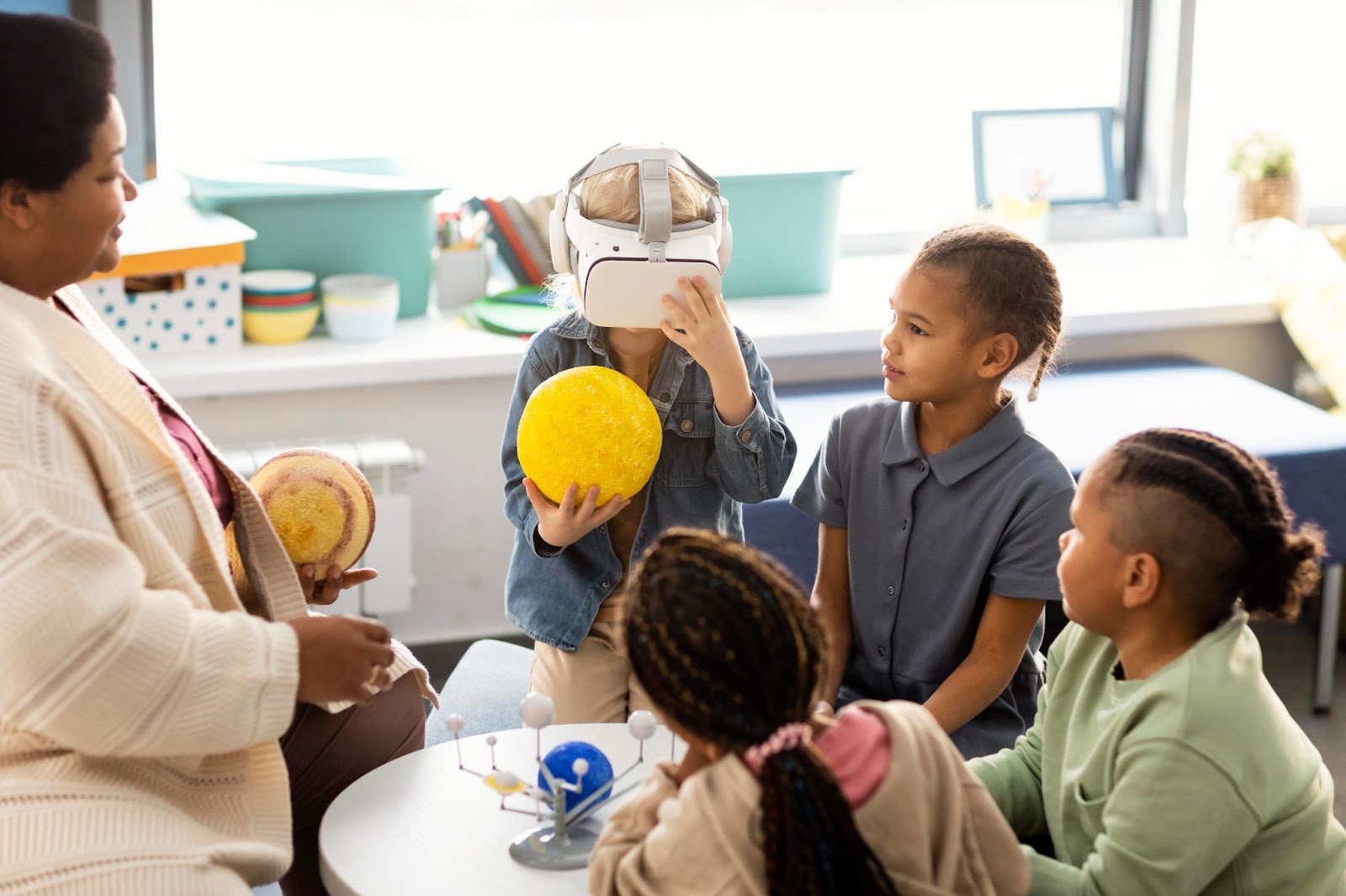 Sala de aula com uma professora e quatro alunos à sua frente, um deles está com óculos de realidade virtual enquanto segura um planeta retirado da representação do sistema solar. A imagem representa o edutainment em sala de aula.