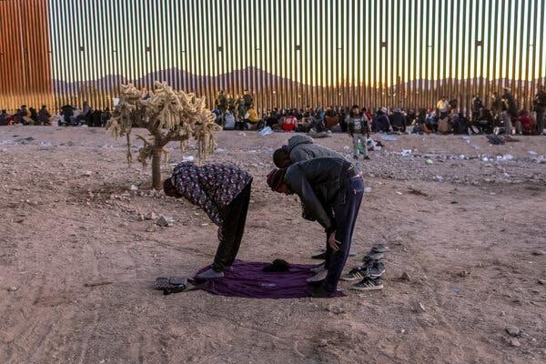 Three people bend over in prayer as they stand on dirt. In the background is a large group of people sitting against a wall. 