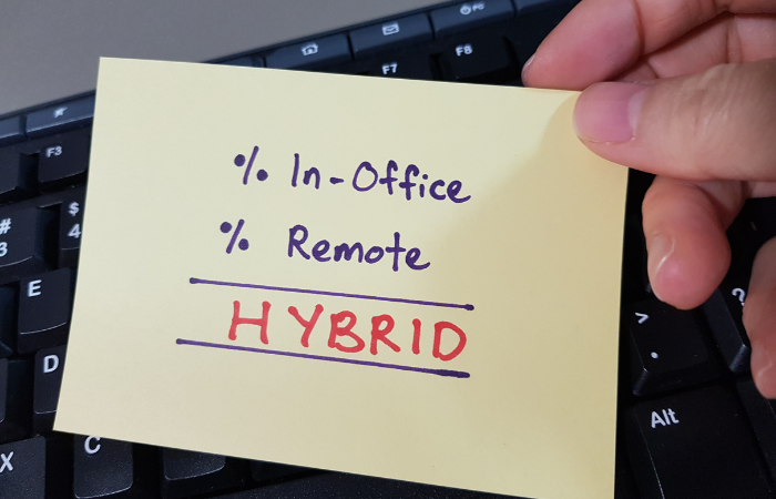 A hand holding a sticky note with "% In-Office % Remote HYBRID" written on it, over a keyboard, indicating a hybrid work model.