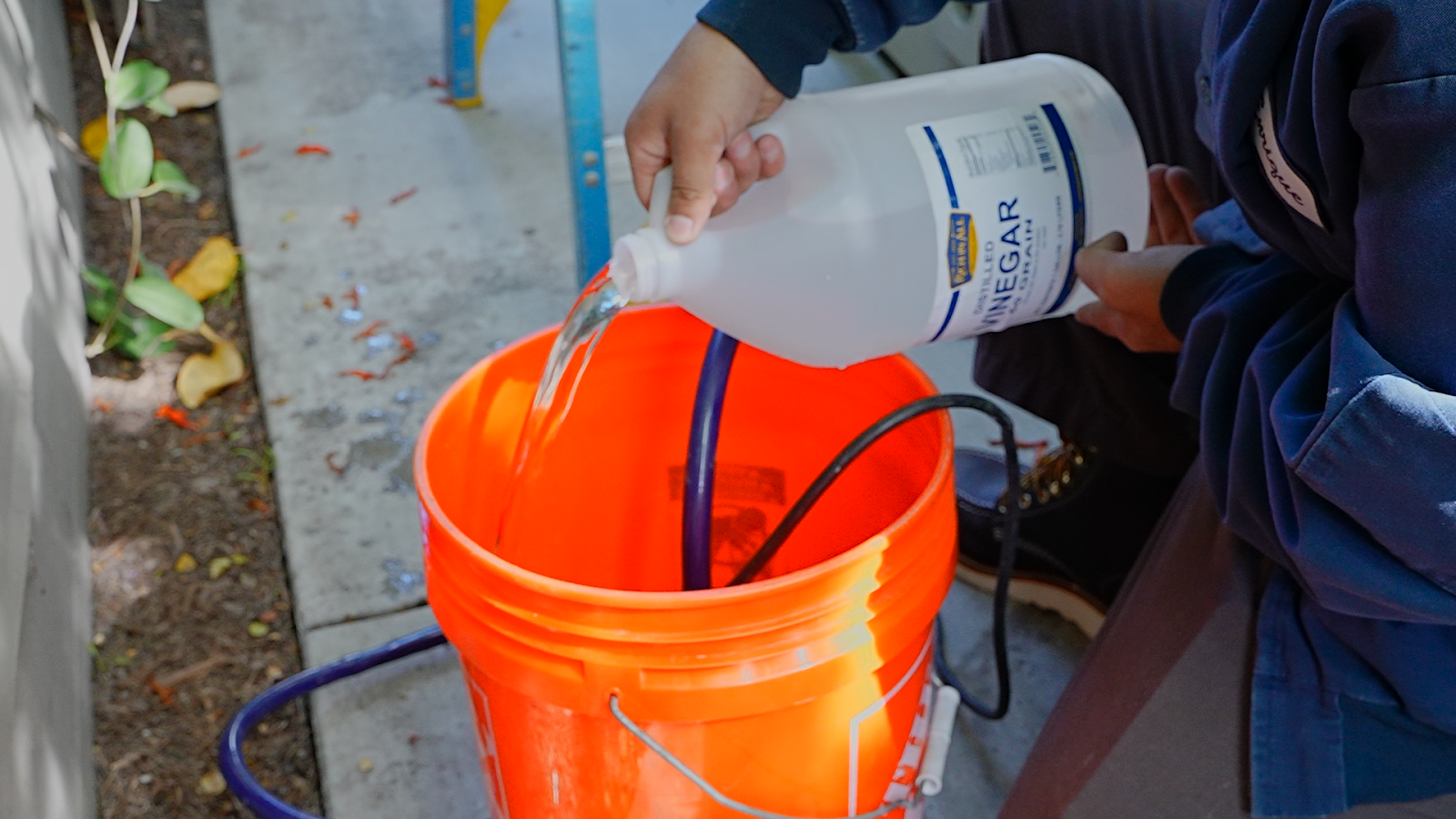 The image shows a Monkey Wrench Plumbing, Heating & Air technician pouring white vinegar into an orange 5-gallon bucket.