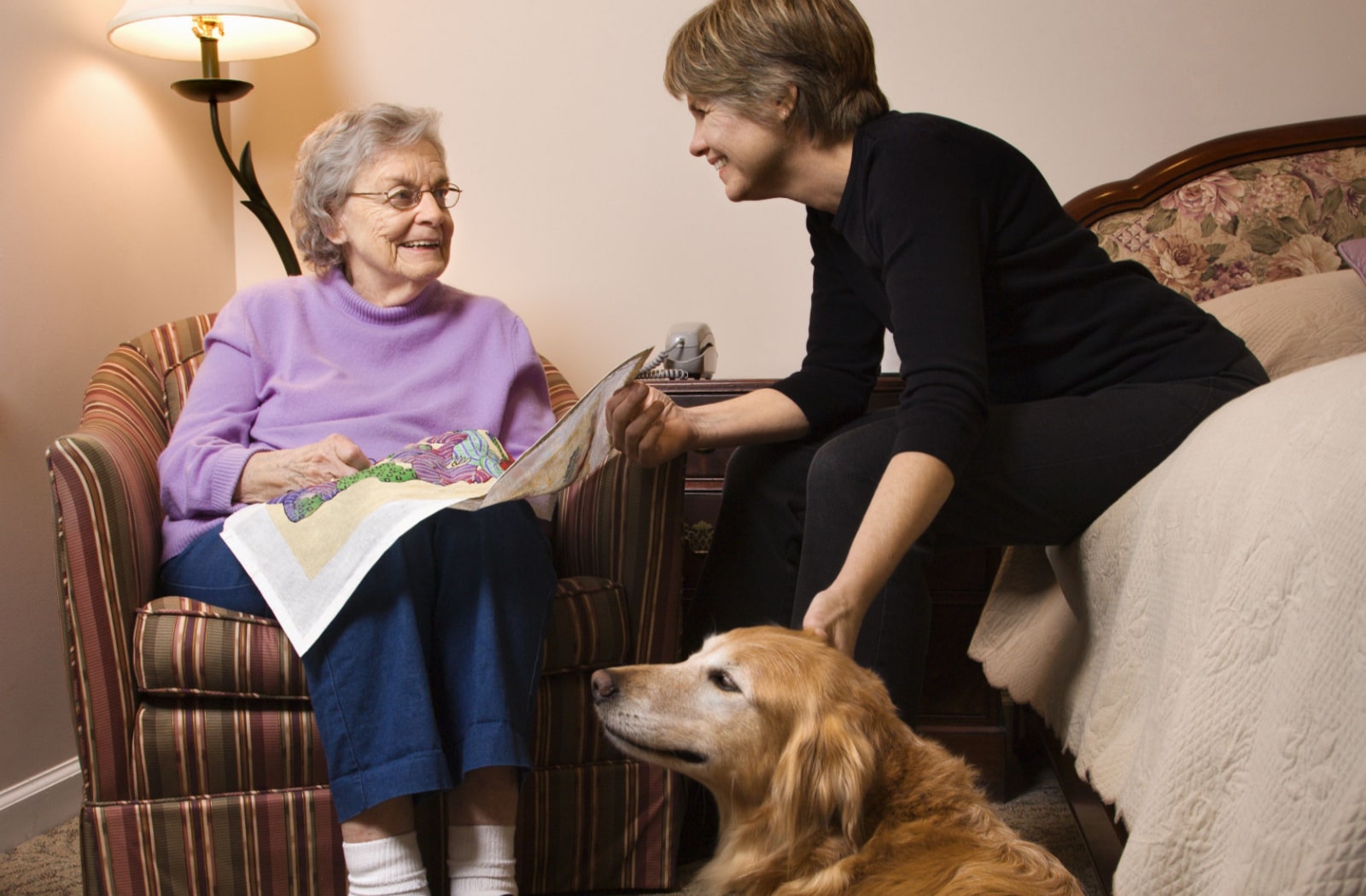 A female dog handler and a female senior living resident sit together in the resident's room and the dog is sitting next to them on the floor