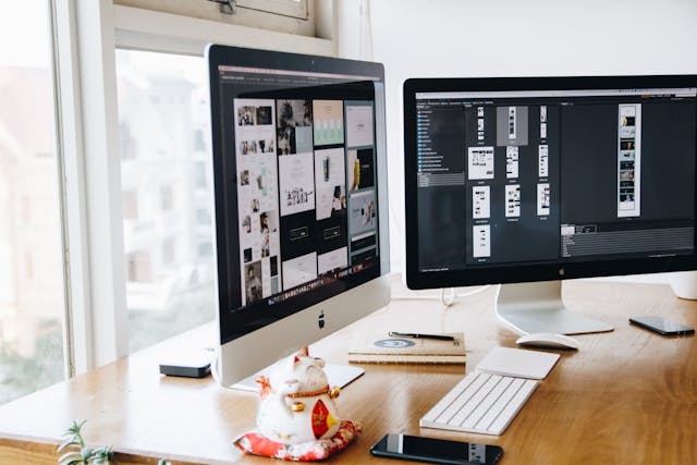 Two monitors with UI UX wireframe facing each other on a wooden desk next to a window