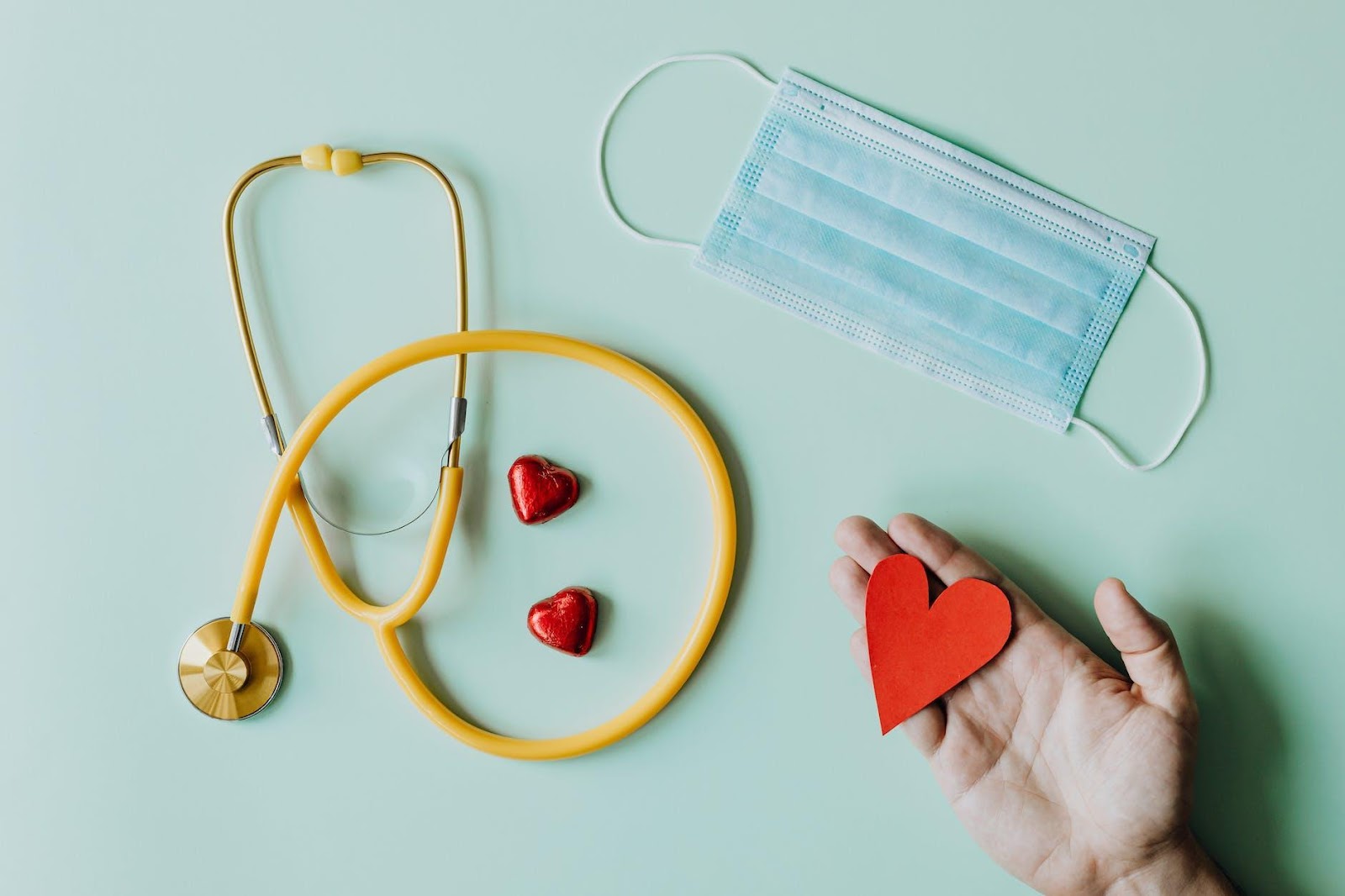 Top view of crop anonymous person hand with red paper heart on table with stethoscope and medical mask