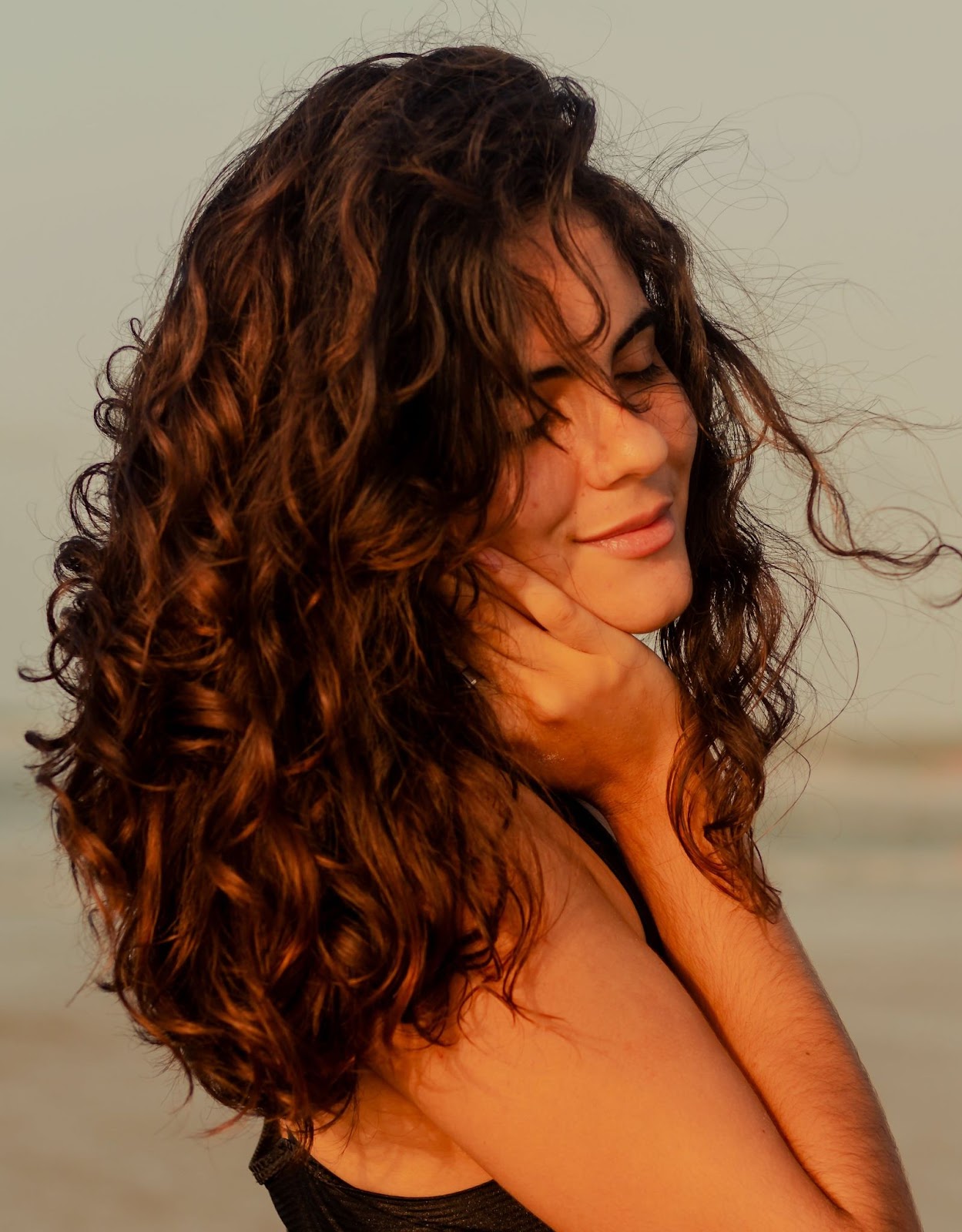 Na imagem, modelo de cabelos ondulados esvoaçantes sorrindo na praia com uma das mãos no rosto