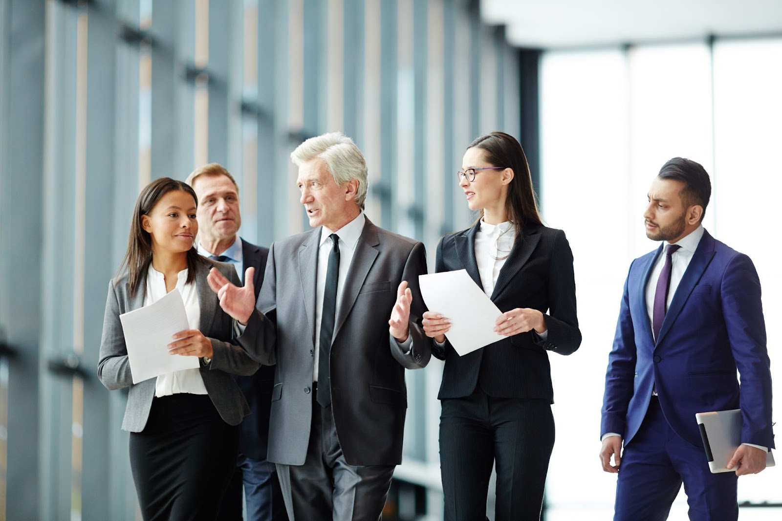 Men and women in suits and holding papers in a glass hallway.