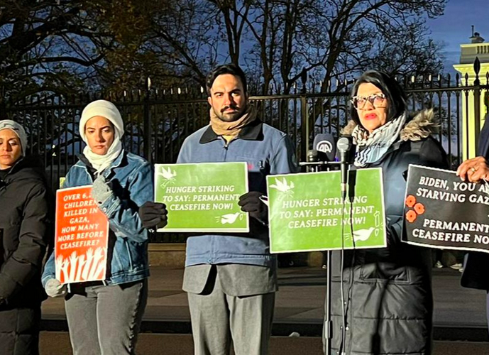 Rashida speaks at a microphone outside the White House at night. She is surrounded by hunger strikers holding signs demanding a permanent ceasefire