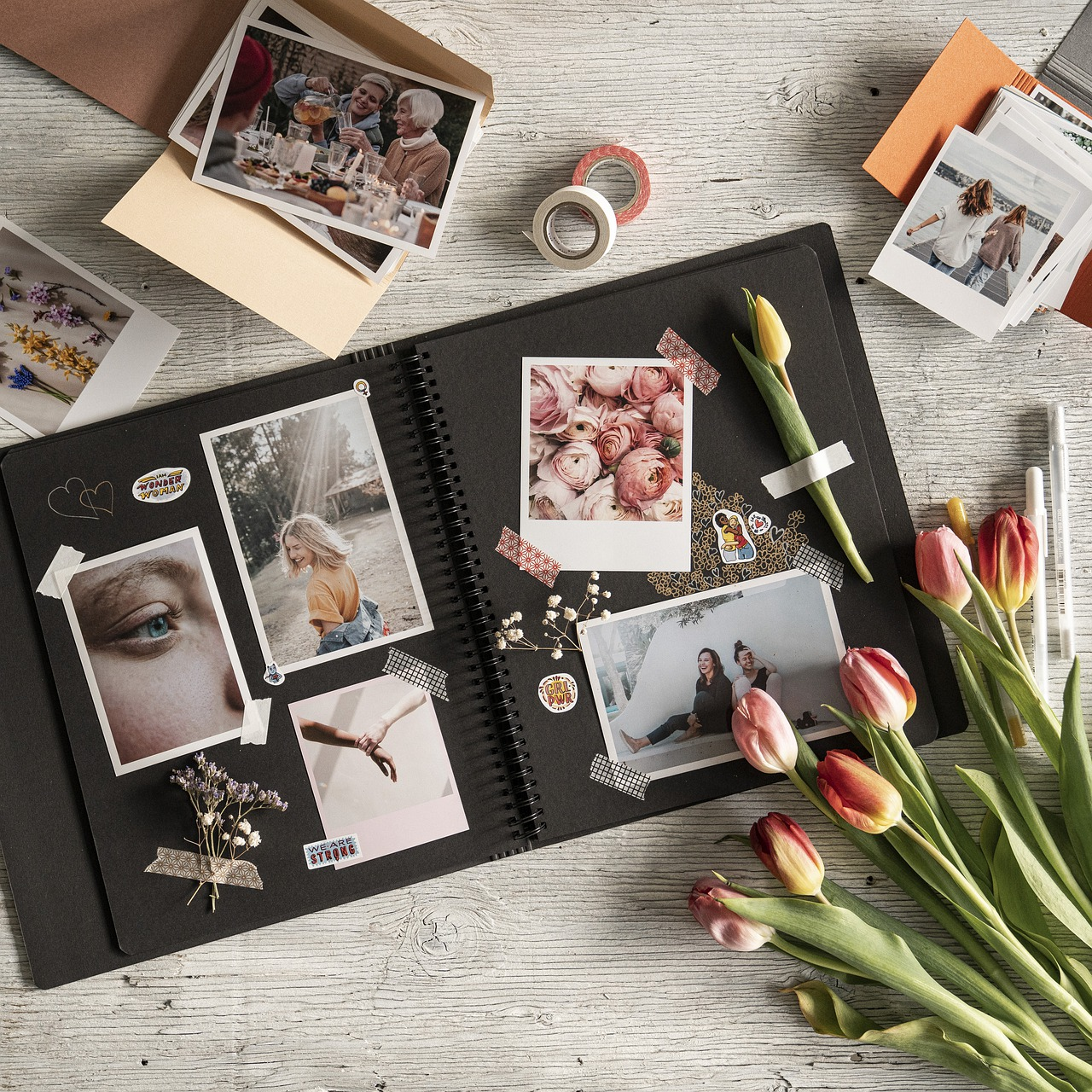 an overhead shot of a dark photo album showing multiple photos next to flowers and rolls of tape