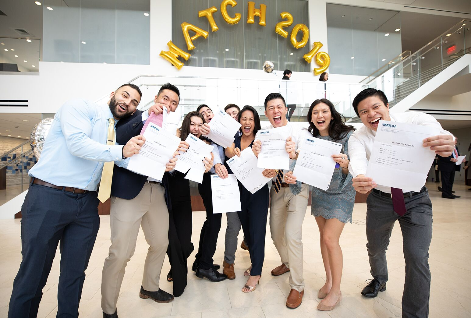 An image of a group of smiling medical students in white lab coats holding envelopes and celebrating together in a room with balloons and decorations. 