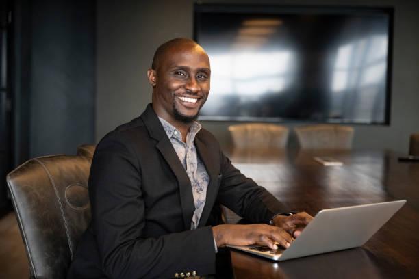 Meeting room portrait of early 30s Black businessman Confident professional wearing blazer over open collar shirt, sitting at conference table, and pausing from typing on laptop to smile at camera. kenyan businessman stock pictures, royalty-free photos & images