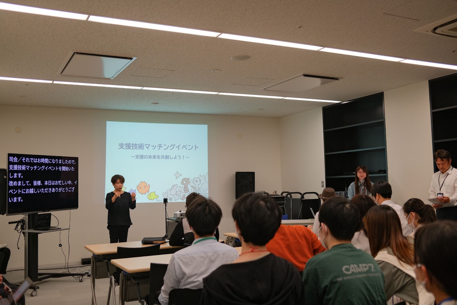 イベント冒頭、筑波大学 担当者がイベント趣旨を説明している。横には、手話通訳士と文字通訳のモニターが並んでいる。