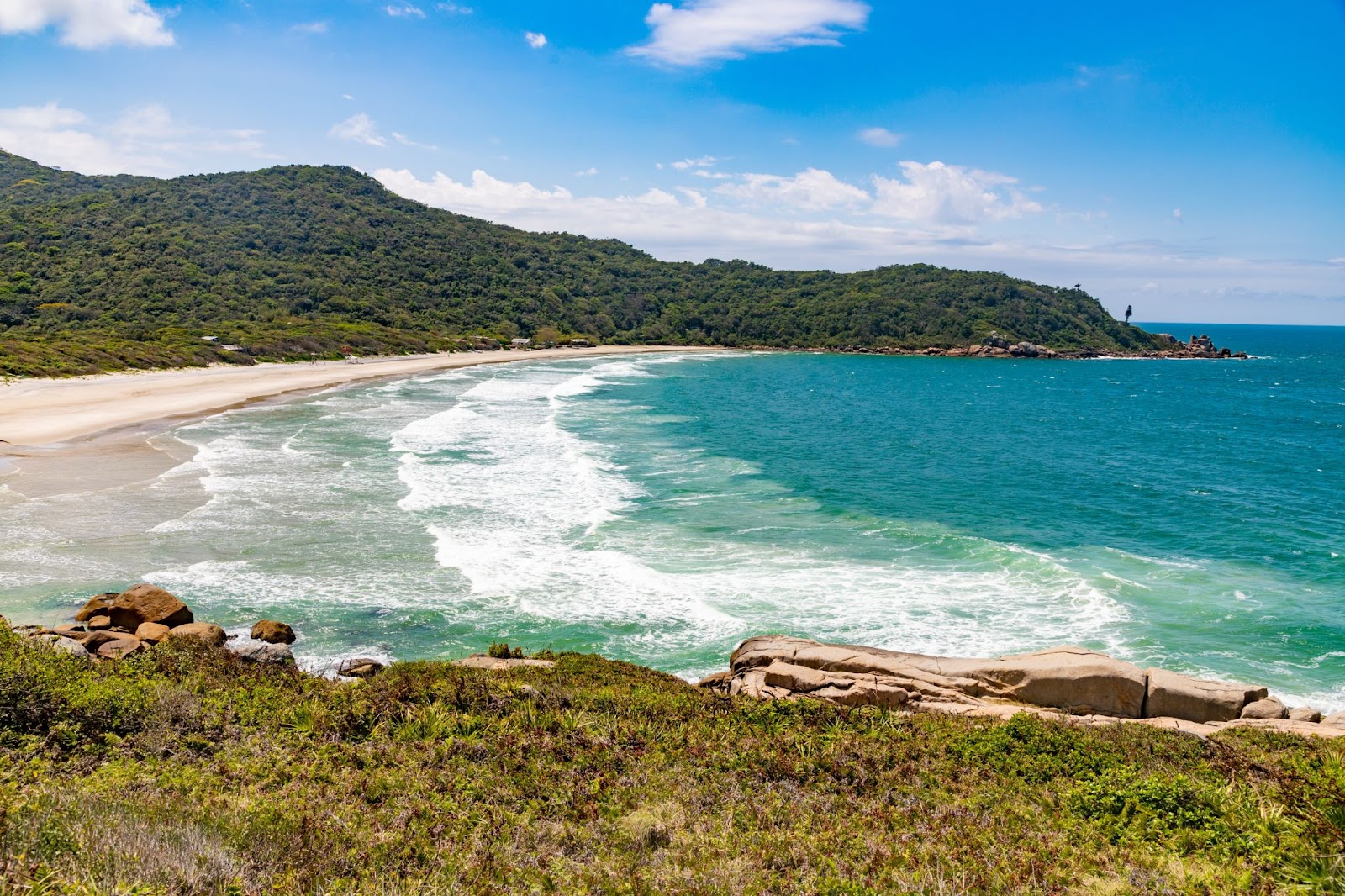 Praia dos Naufragados, em Florianópolis. O mar de coloração azul intensa forma ondas muito leves e quebra em uma fina faixa de areia. Ao fundo, aparecem montanhas com vegetação densa e verde.