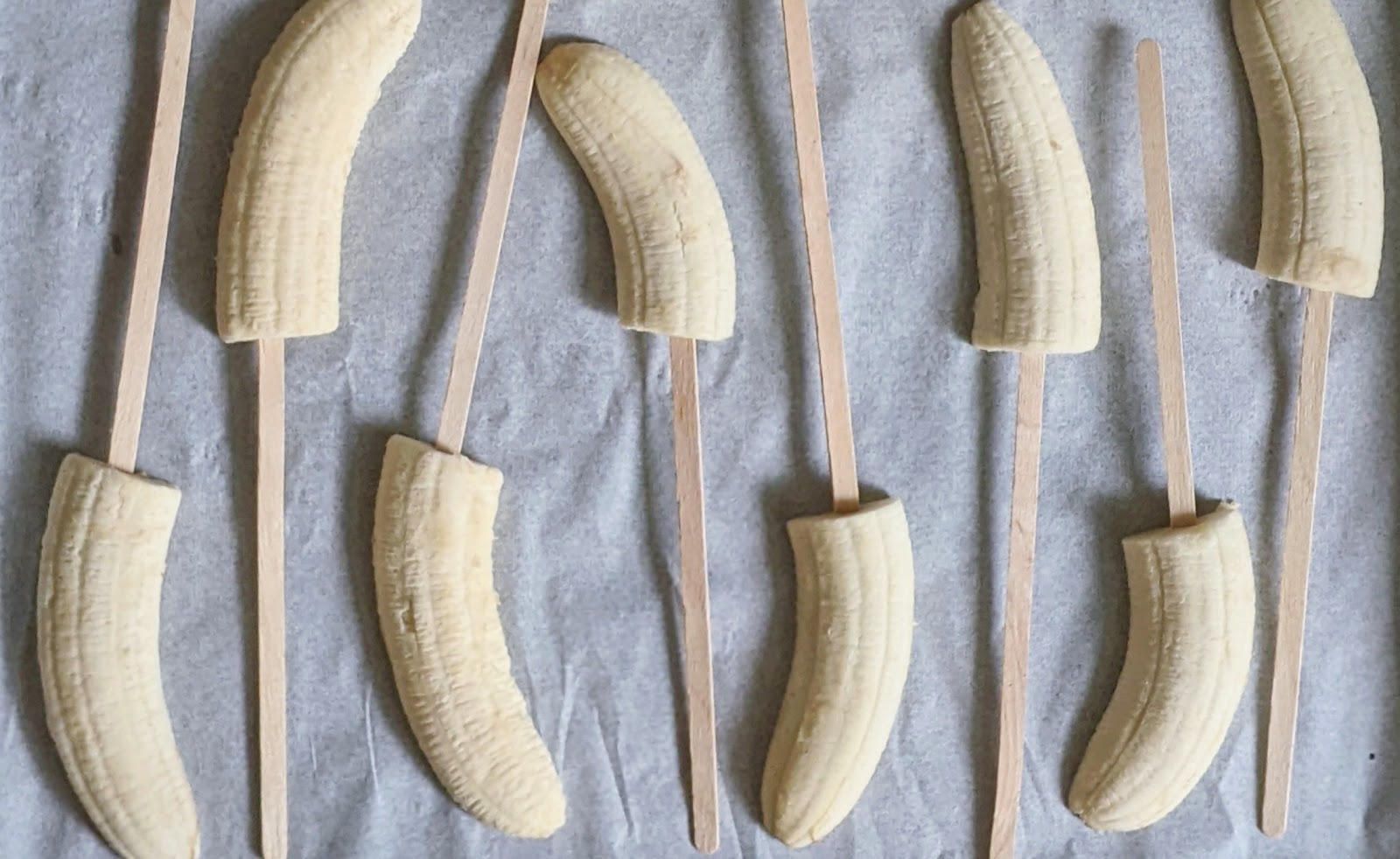 top down view of sliced bananas with wooden sticks inserted into them laying on a baking tray with parchment paper