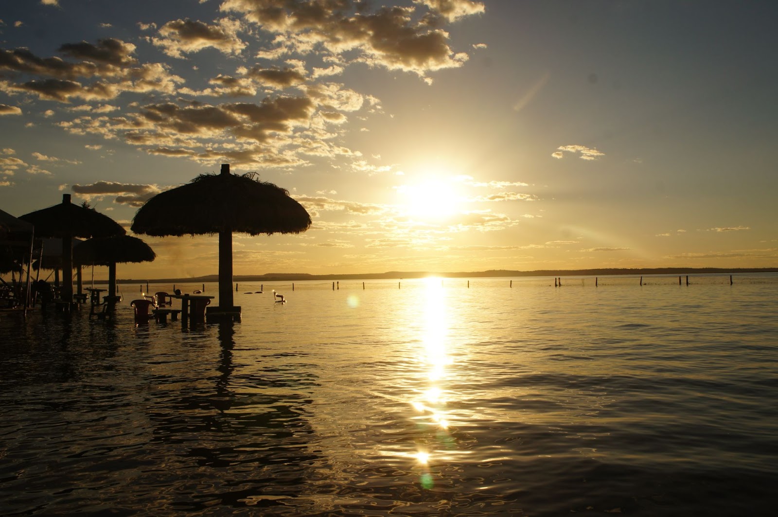 Foto do pôr do sol na Ilha do Canela. Ao lado esquerdo, guarda-sóis feitos de palha, com banquinhos na água