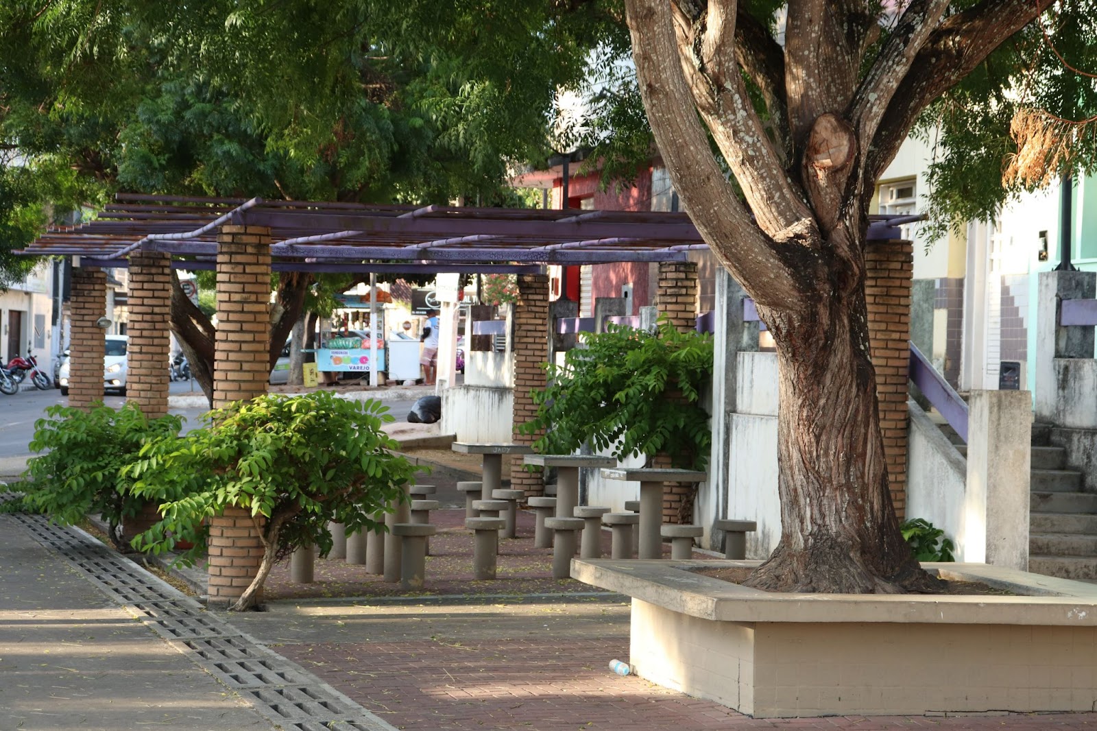 Foto de uma das praças da cidade de Arapiraca. Diversas mesas e bancos de concreto embaixo de uma estrutura para proteger do sol