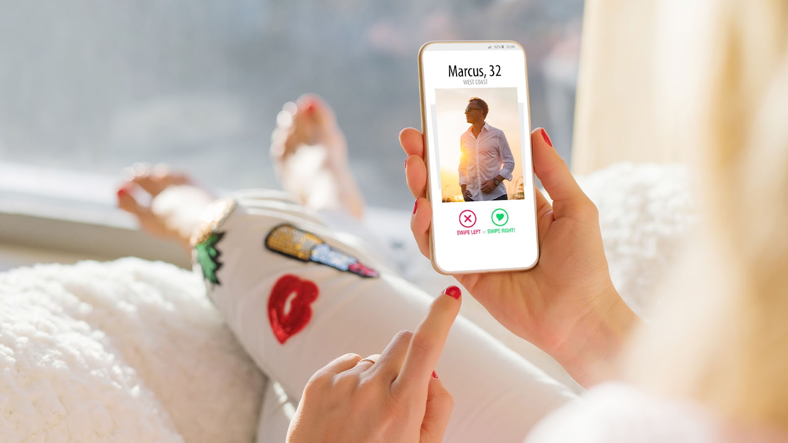 Blonde woman in pyjamas lying on a pillow holding her phone which shows the the dating profile of a man