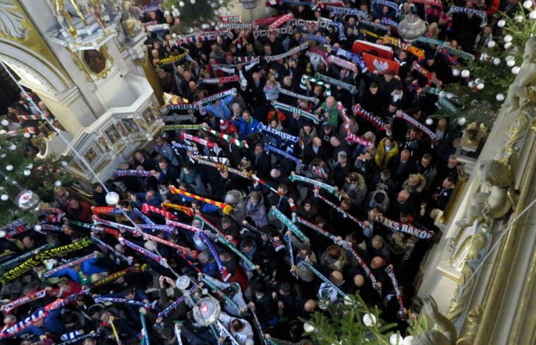 Zdjęcie Kibiców w kościele. Kibice trzymają górze szaliki - barwy swoich klubów.