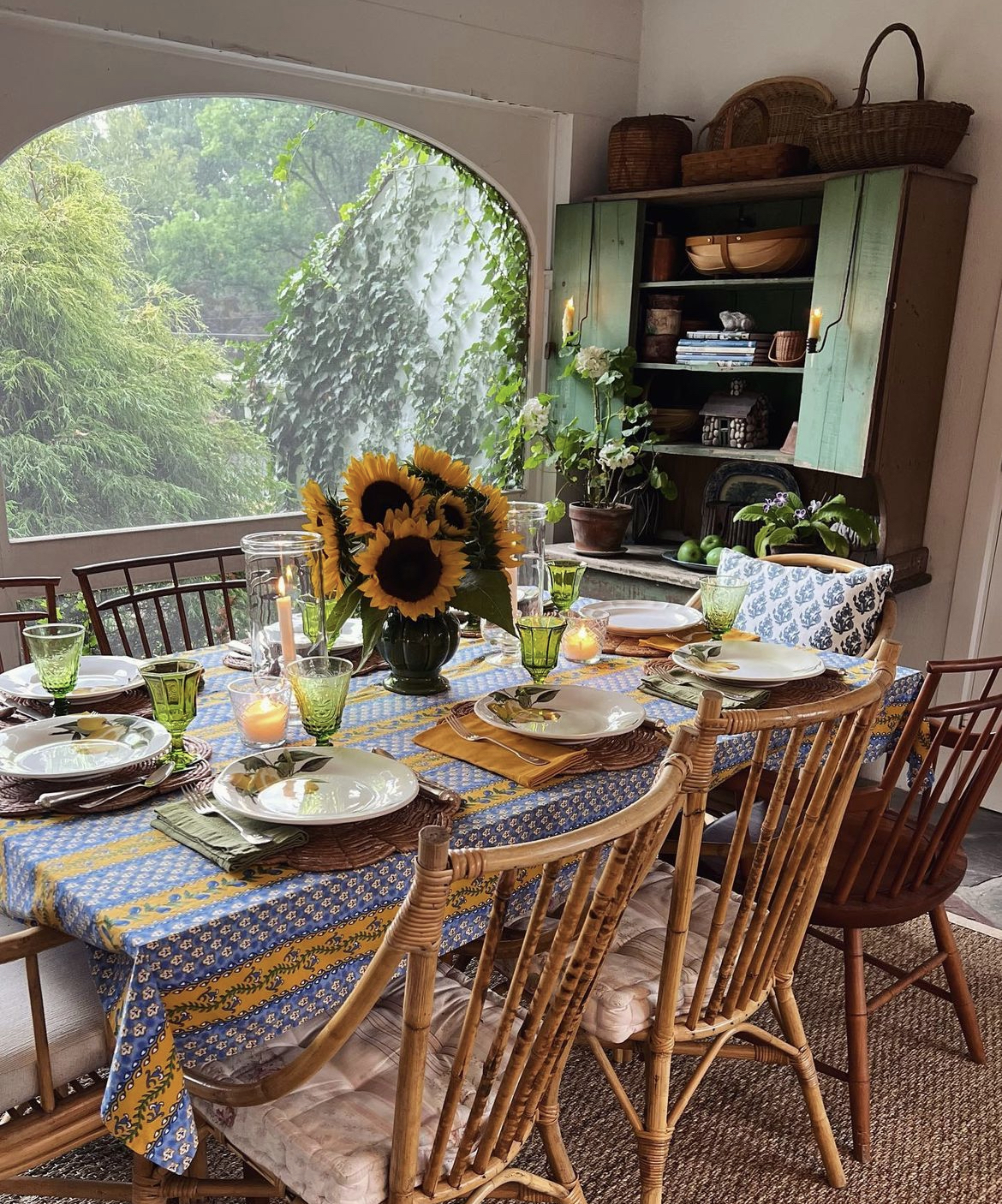 A cottage-inspired tablescape featuring a mix of vintage chairs and colorful place settings upon a blue and gold patterned tablecloth centered by a bouquet of sunflowers.