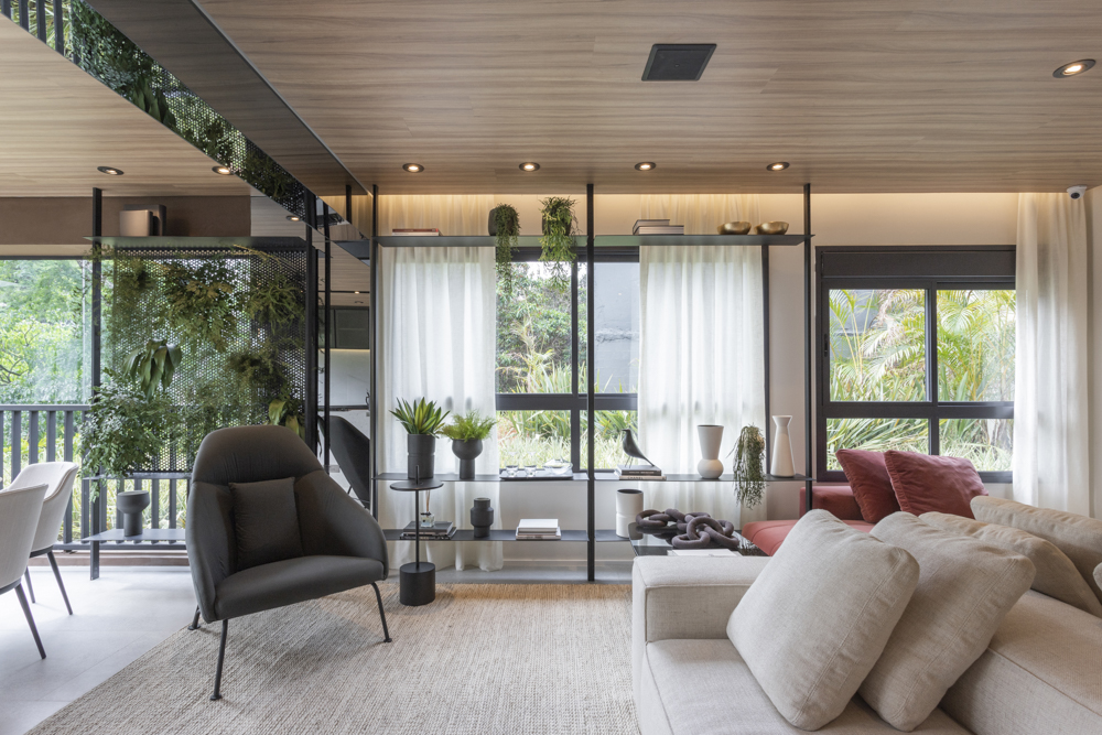 Sala de estar com um lindo jardim vertical próximo a janela com cortinas brancas e sofá na cor bege e poltrona escura.