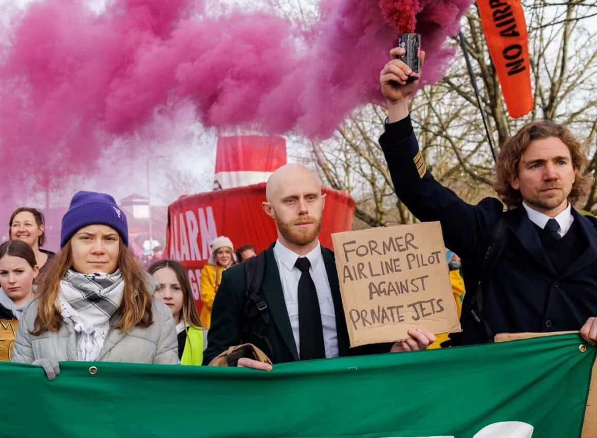 Greta marches beside two uniformed activists - one holding a sign saying 'former airline pilot against private jets'.