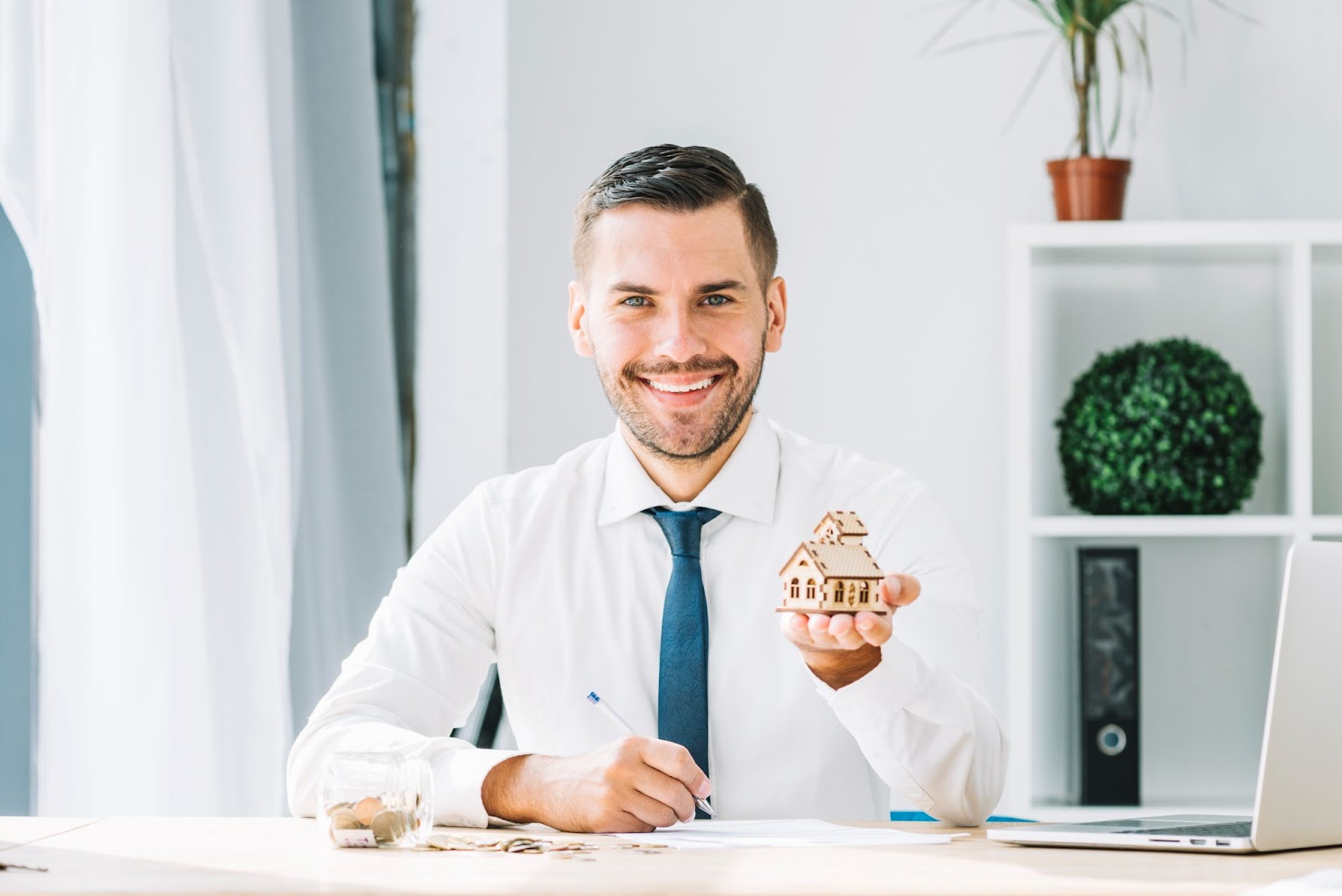 A realtor in a white shirt with a blue tie is posing with a miniature house in his hand to convey his message to potential clients. 