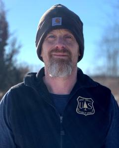 Outdoor portrait of a man wearing a blue forest service vest, long sleeves and a knit hat. His goatee is red and grey.