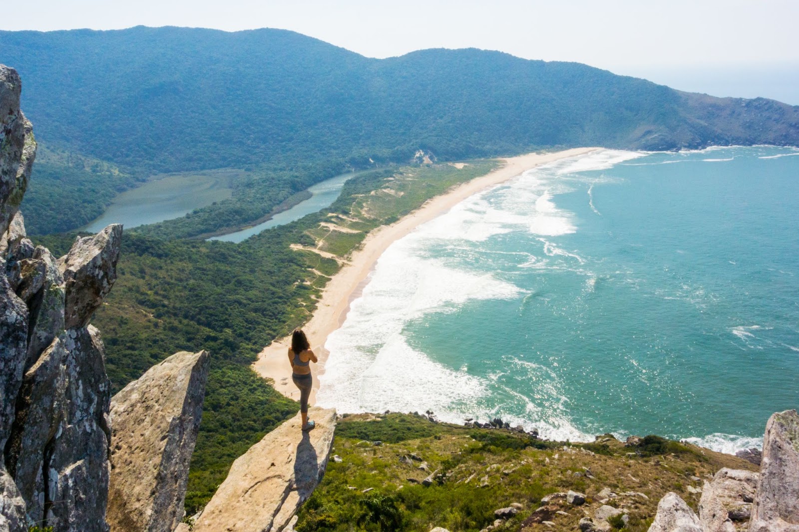 Turista de pé em uma pedra suspensa alta, no Morro da Coroa. Ela contempla o mar claro da Praia da Lagoinha do Leste, que aparece embaixo, e as montanhas verdes.