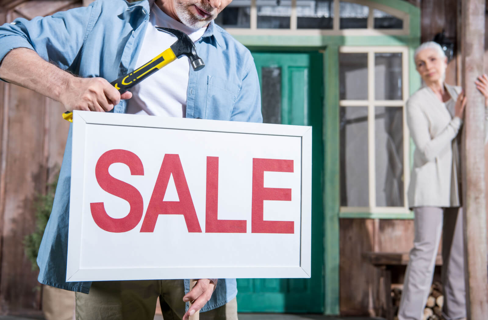 An older adult man putting a sale sign on their house with his wife in the background.